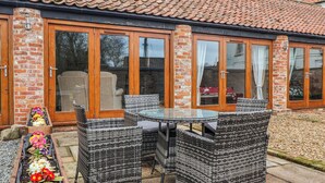 A patio with wicker furniture, a glass table, and a flower bed with various flowers. - Pillar Barn (Driffield)