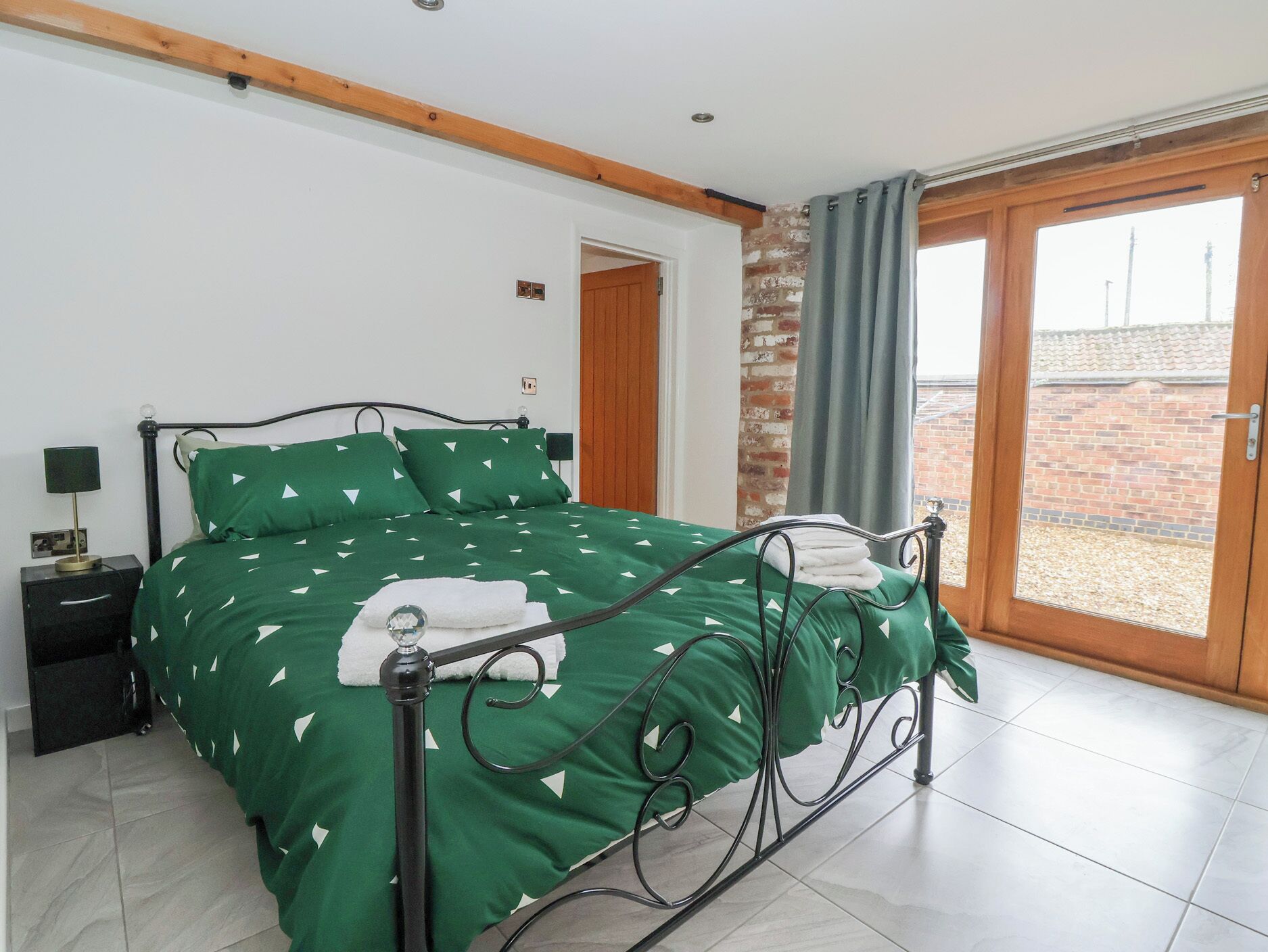 Un dormitorio con cubrecama verde, estructura de cama metálica, mesita de noche con lámpara y vista a una pared de ladrillo a través de una puerta corrediza.