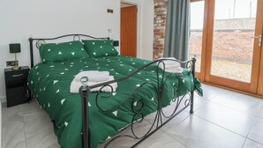 A bedroom with a green bedspread, a metal bed frame, a nightstand with a lamp, and a view of a brick wall through a sliding door. - Pillar Barn (Driffield)