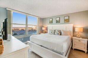 A bedroom with a large bed, two bedside tables with lamps, a TV on a stand, and a view of the city through large windows. - Magnolia House at Destin Pointe 203 (Destin)