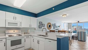 A modern kitchen with white cabinets, a microwave, an oven, and a dishwasher. There is a dining area with a table and chairs, and a view of the outside. - Coastal condo with Gulf views, washer and dryer, Wifi, Community pool (Destin)