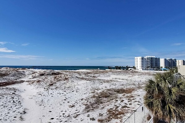 A beach with sandy terrain, a clear blue sky, and a few buildings in the background.
