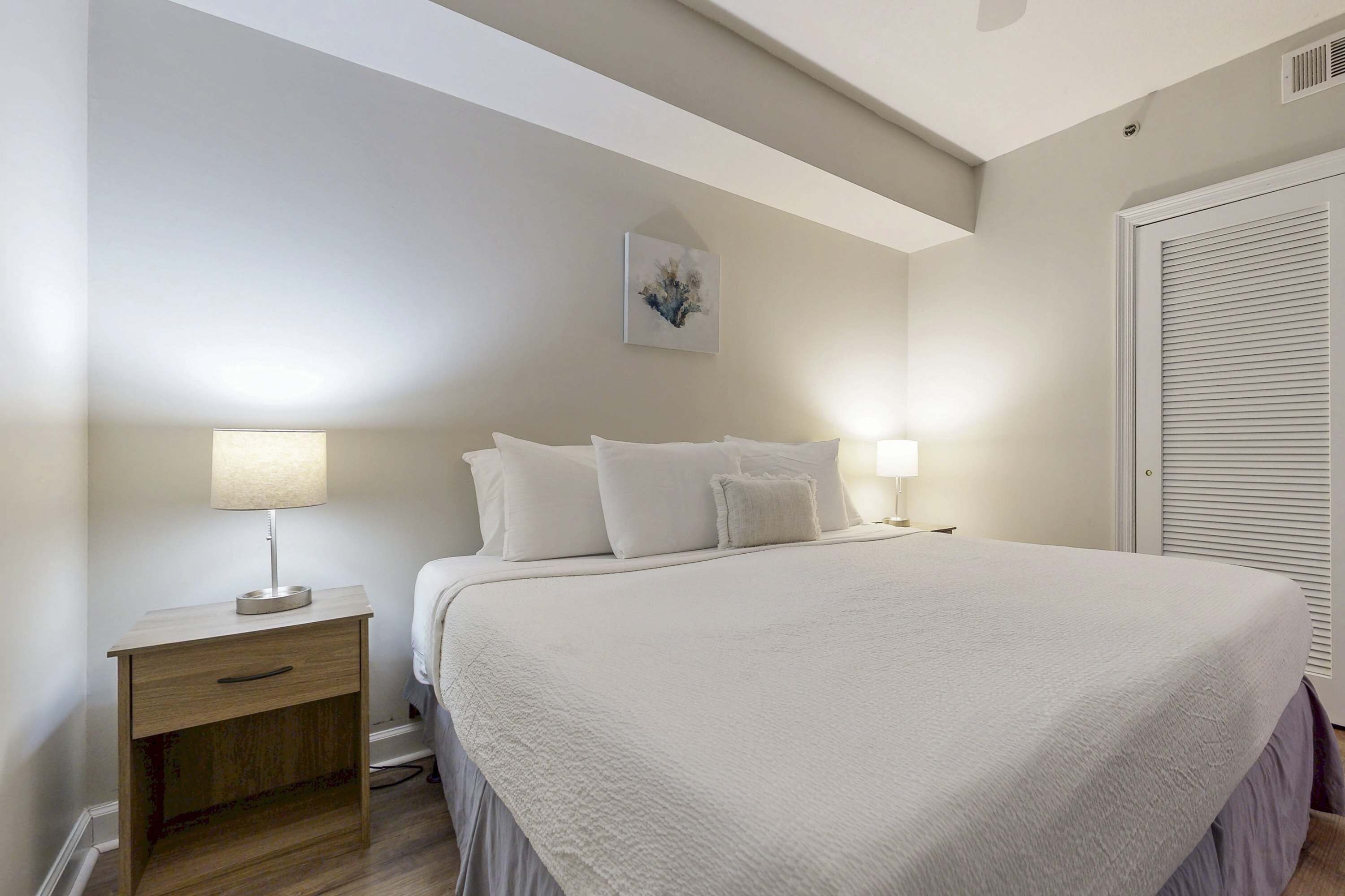 A bedroom with a large bed, two bedside tables with lamps, a nightstand, a ceiling light, and a door with a louvered design.