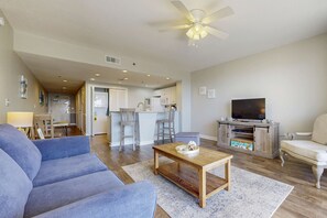 A living room with a sofa, coffee table, TV, and a kitchen in the background. - Ocean-View Getaway with Two Shared Pools, Gym & Beach Access (Destin)