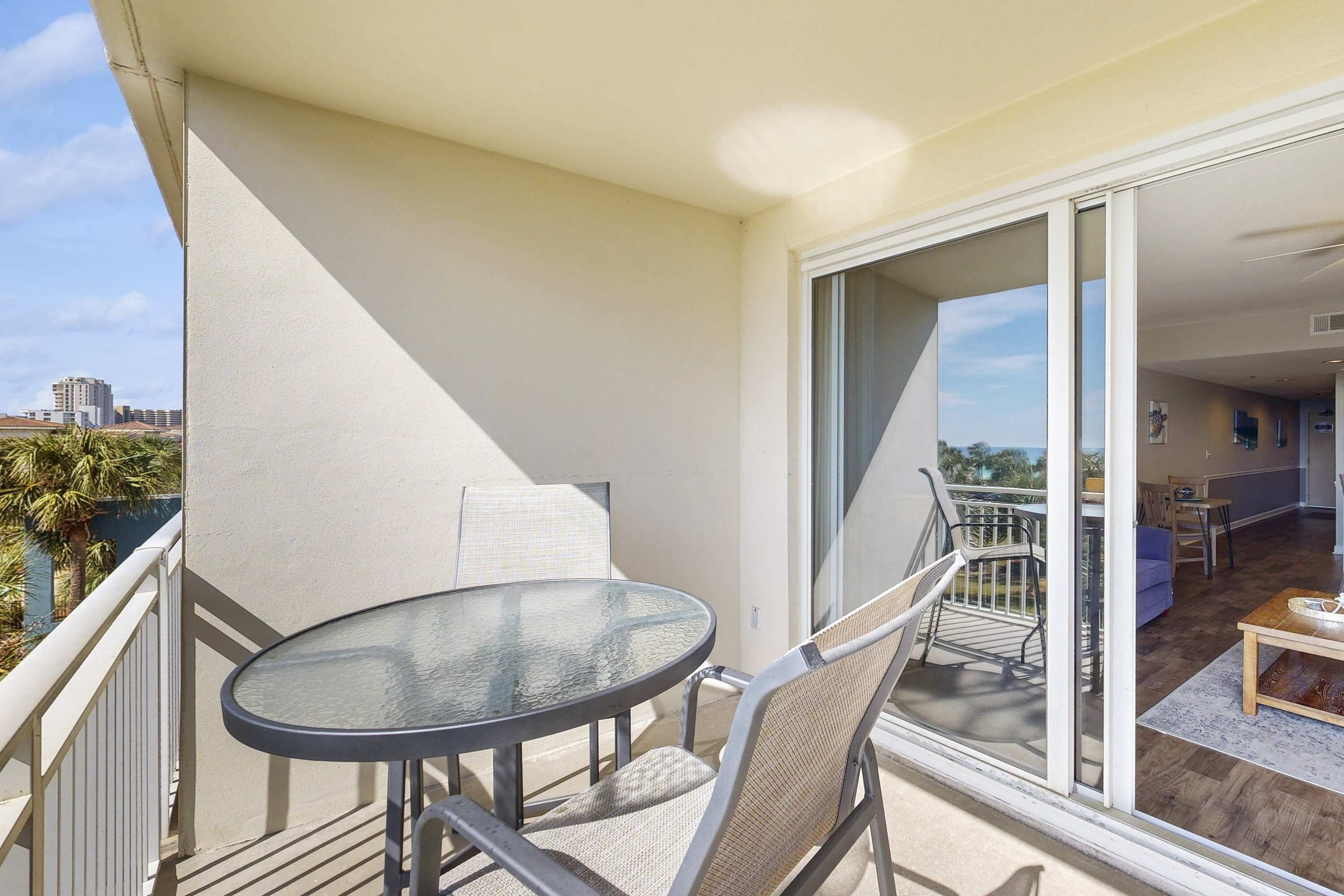A balcony with a glass table and chairs, a sliding glass door leading to an interior space with a sofa and a coffee table.
