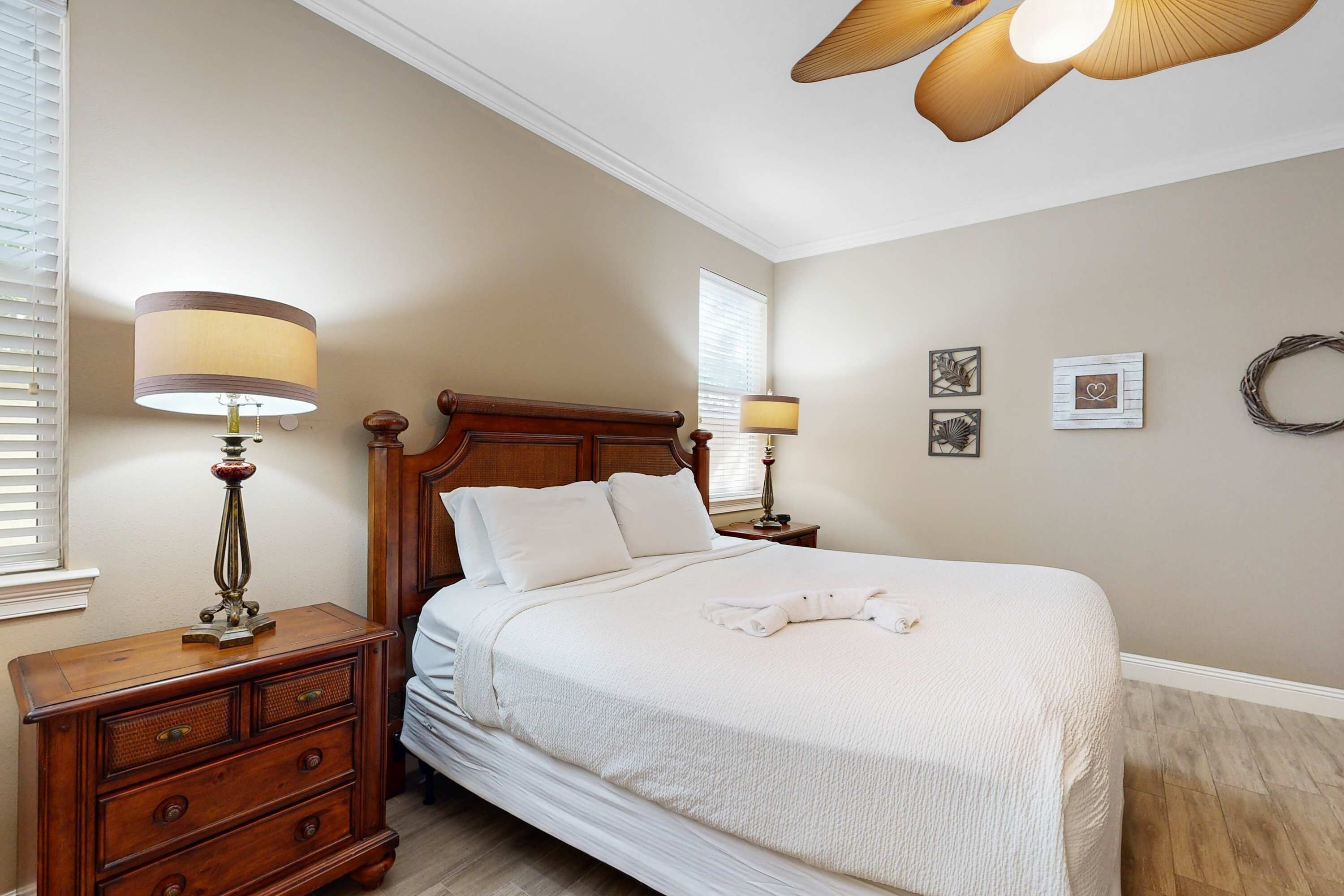 Une chambre à coucher avec un grand lit, une tête de lit en bois, deux tables de chevet avec des lampes, une commode et un ventilateur de plafond.
