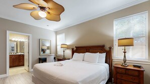 A bedroom with a large bed, a wooden nightstand with a lamp, a ceiling fan, and a window with blinds. - Nautical home in gated community with pool, hot tub & beach access (Destin)