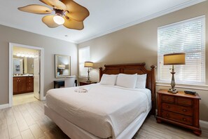 A bedroom with a large bed, a wooden nightstand with a lamp, a ceiling fan, and a window with blinds. - Nautical home in gated community with pool, hot tub & beach access (Destin)