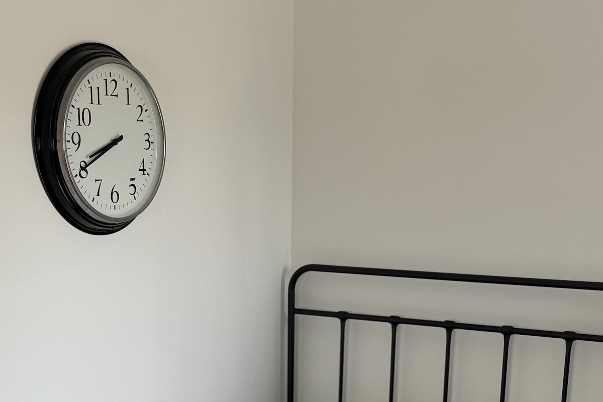 A single bed with a metal frame, a grey patterned bedspread, two brown pillows, and a white clock on the wall.