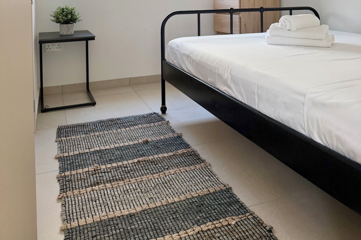 A bedroom with a bed, a bedside table, a window with a blind, a small rug, and a picture frame on the wall.