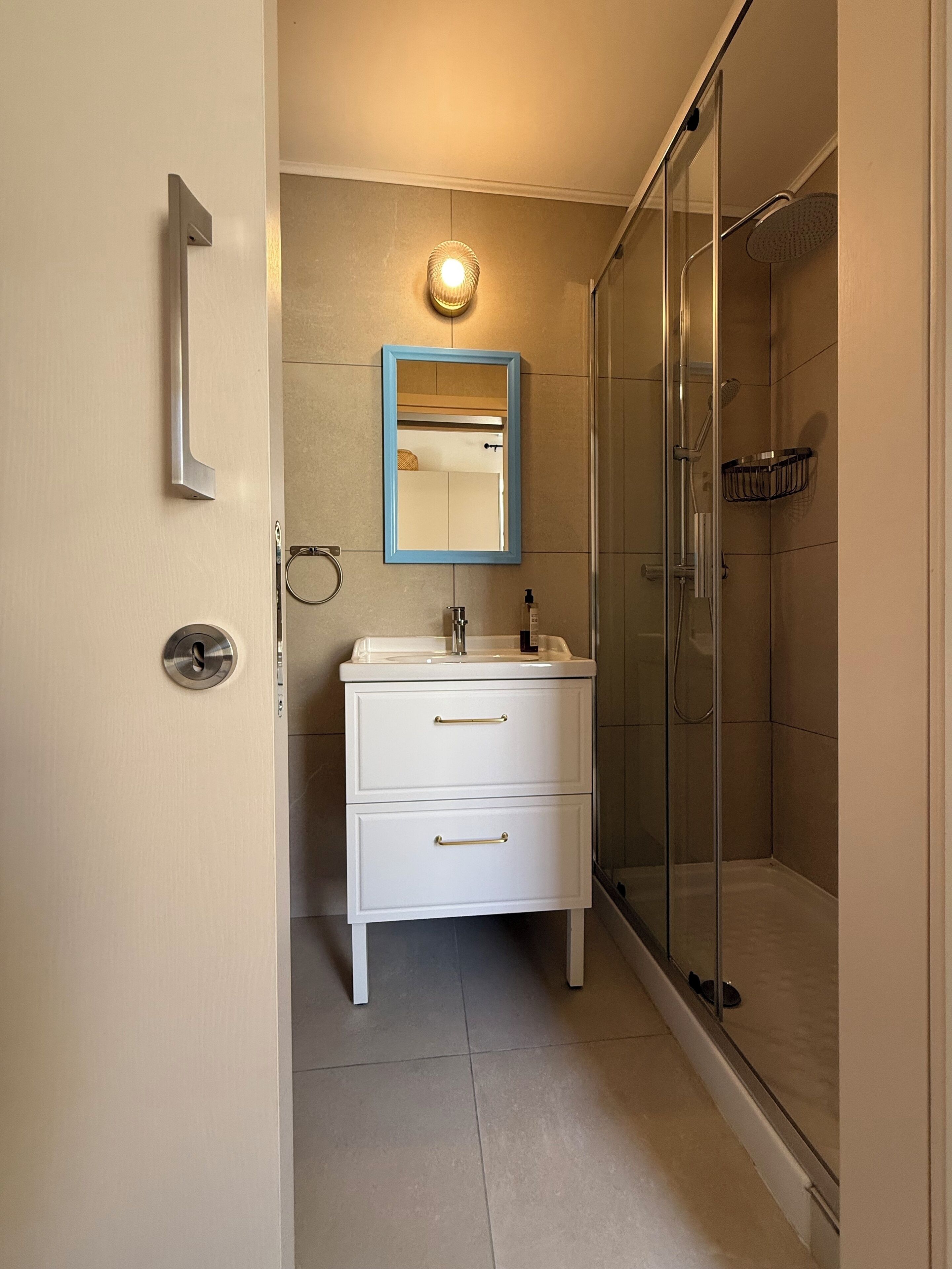 Een badkamer met een douchecabine van glas, een witte wastafel met lades, een spiegel met blauwe lijst en een wandlamp.
