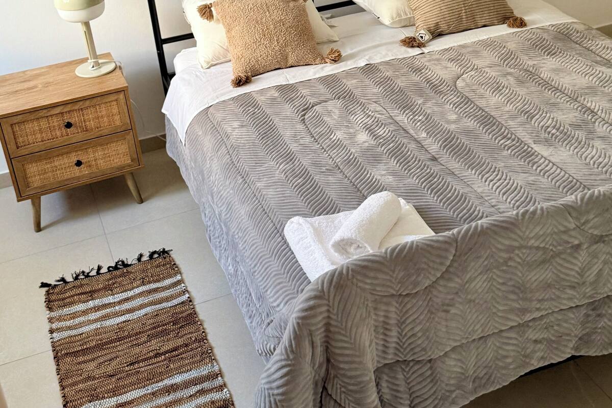 A neatly made bed with a striped blanket, a bedside lamp, a woven rug, and a wooden nightstand.