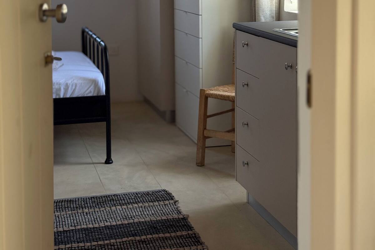A narrow hallway with a bed, a wooden stool, a dresser, and a plant.
