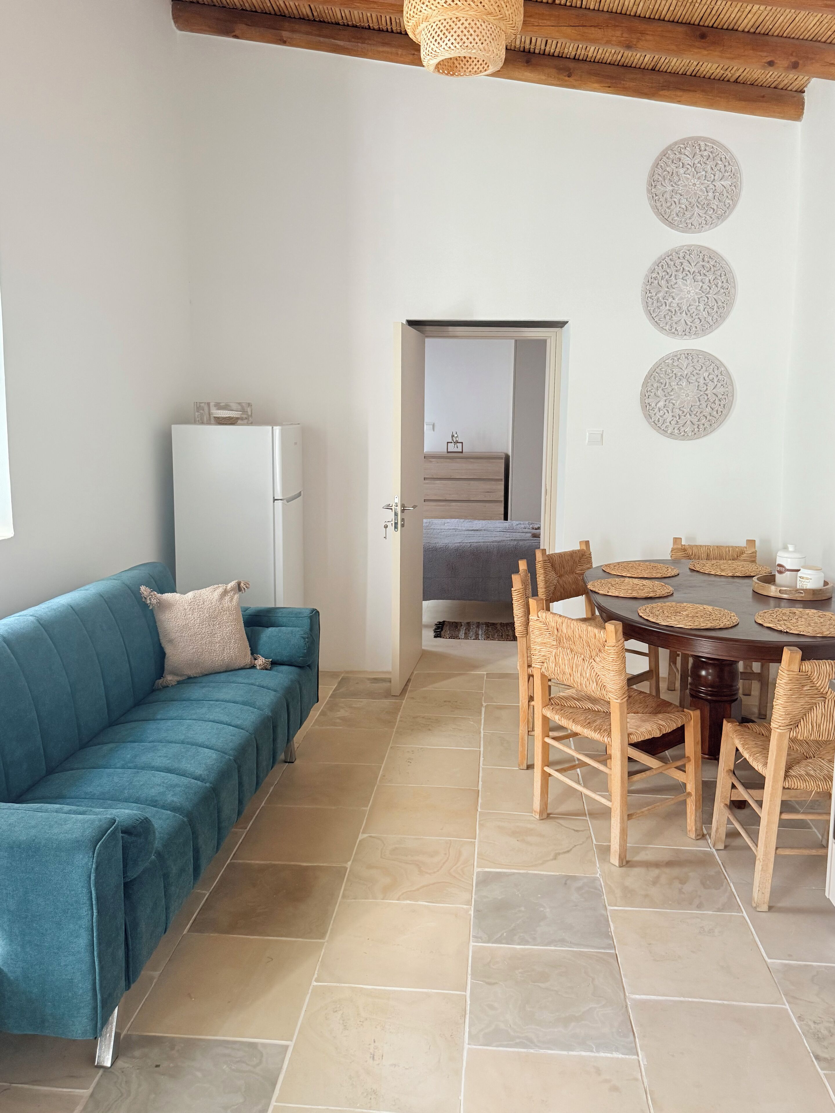 Ein modernes Wohnzimmer mit einem petrolfarbenen Sofa, einem Esstisch mit Korbstühlen und einem weißen Kühlschrank.