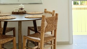A dining area with a round table, four wicker chairs, and a window with a view of buildings and a clear sky. - Kiti Traditional Residences (Kiti)
