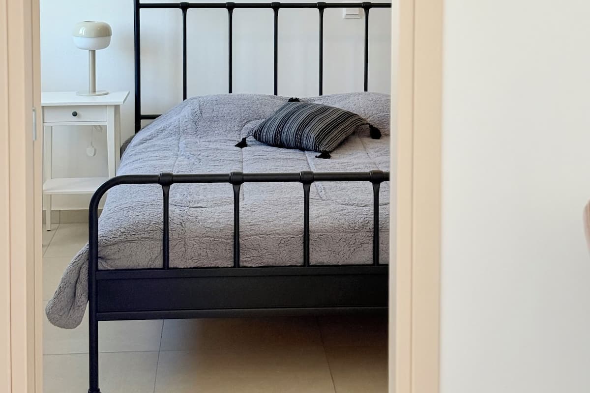 A bed with a metal frame and a grey textured mattress, a bedside table with a lamp, and a white wall.