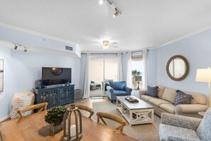 A living room with a television, sofa, coffee table, and dining table. - Lovely waterfront condo with balcony, pool, hot tub & pickleball (Santa Rosa Beach)