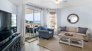 A living room with a sofa, coffee table, and TV. There is a balcony with a view of the ocean. - Lovely waterfront condo with balcony, pool, hot tub & pickleball (Santa Rosa Beach)