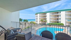 A balcony with wicker furniture, a pool, and a view of a beach and hotel building. - Lovely waterfront condo with balcony, pool, hot tub & pickleball (Santa Rosa Beach)