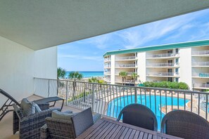 A balcony with wicker furniture, a pool, and a view of a beach and hotel building. - Lovely waterfront condo with balcony, pool, hot tub & pickleball (Santa Rosa Beach)