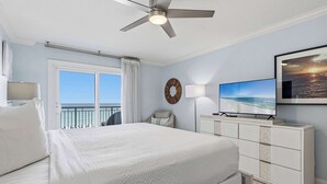 Een hotelkamer met een bed, een plafondventilator, een televisie, een dressoir, een raam met gordijnen en uitzicht op zee.