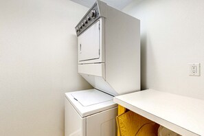A compact laundry room with a stacked washer and dryer, a white countertop, and a wooden cabinet below. - Gulf-front 3-bedroom condo with balconies off each bedroom & pickleball (Santa Rosa Beach)