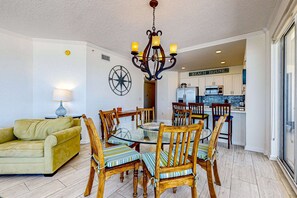 A dining area with a glass table surrounded by wooden chairs, a chandelier, a sofa, and a kitchen in the background. - Gulf-front 3-bedroom condo with balconies off each bedroom & pickleball (Santa Rosa Beach)