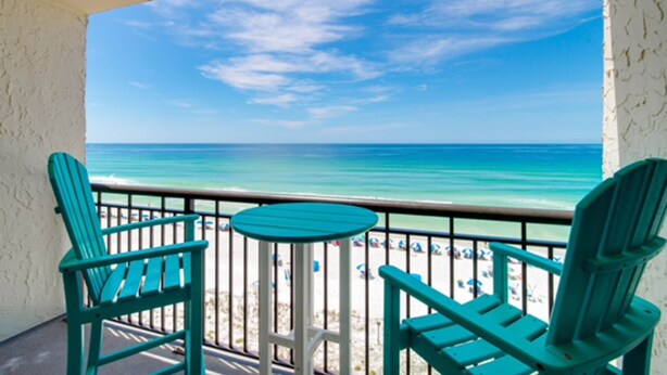 Ein Balkon mit türkisfarbenen Stühlen und einem kleinen Tisch, mit Blick auf einen Strand und einen wolkenlosen, tiefblauen Himmel sowie das türkisfarbene Meer.