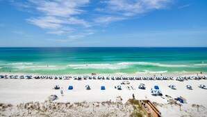 Ein Strand mit Reihen blauer Sonnenschirme und Liegestühle, ein sandiger Uferbereich und das Meer im Hintergrund.