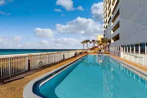 A pool area with a view of the ocean, a white building with balconies, and palm trees. - Top-floor penthouse condo with amazing view, balcony, W/D, pools, gym, & hot tub (Panama City Beach)