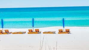 Ein Strand mit Reihen aus Holzliegestühlen und blauen Sonnenschirmen, die entlang der Küste angeordnet sind.