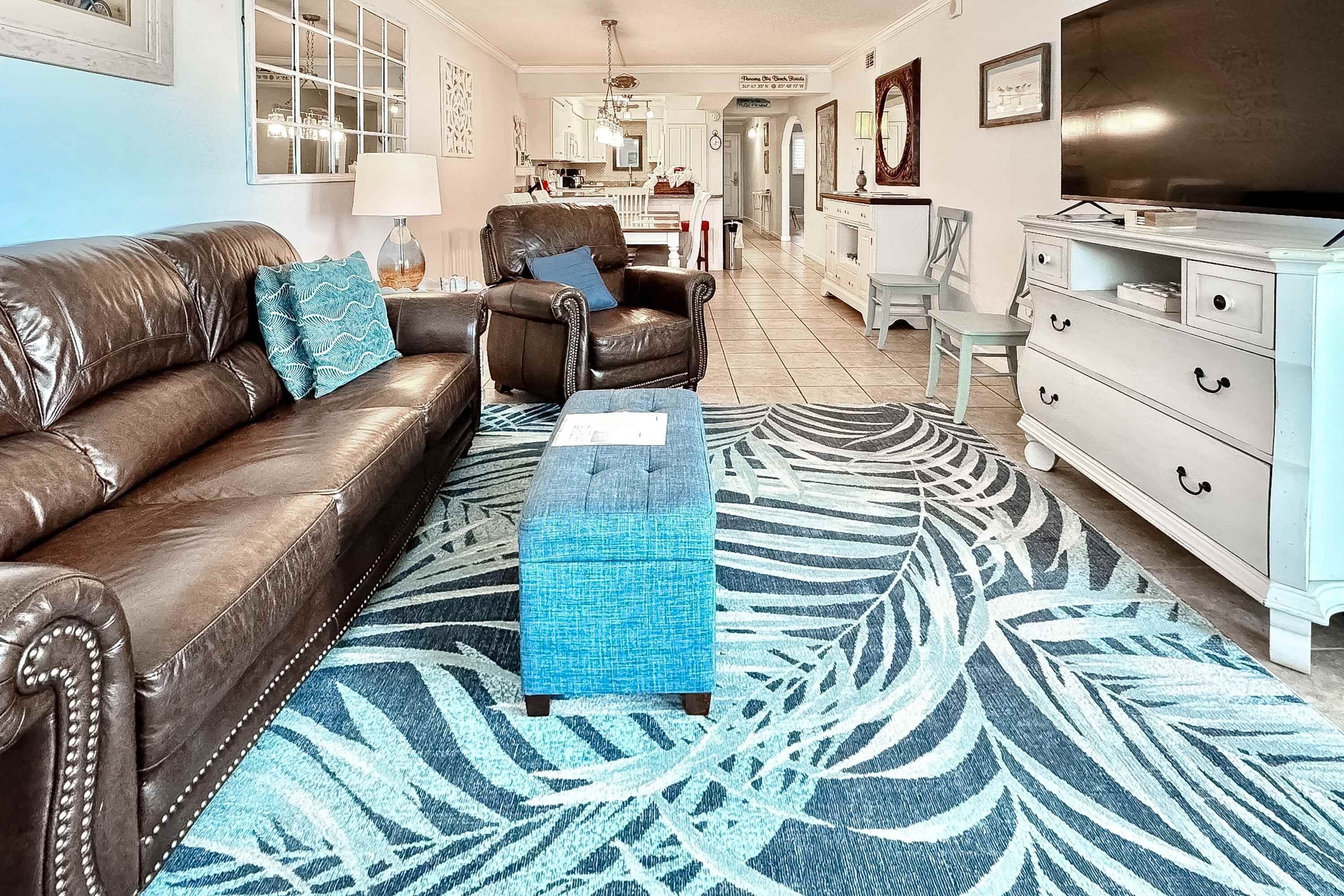 Uma sala de estar com um sofá de couro marrom, um banquinho azul e um tapete estampado.
