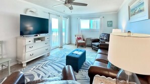 Un salon doté d’un téléviseur à écran plat, d’un ventilateur de plafond, d’une commode blanche, d’un pouf bleu, de canapés en cuir et d’une grande fenêtre avec des rideaux.