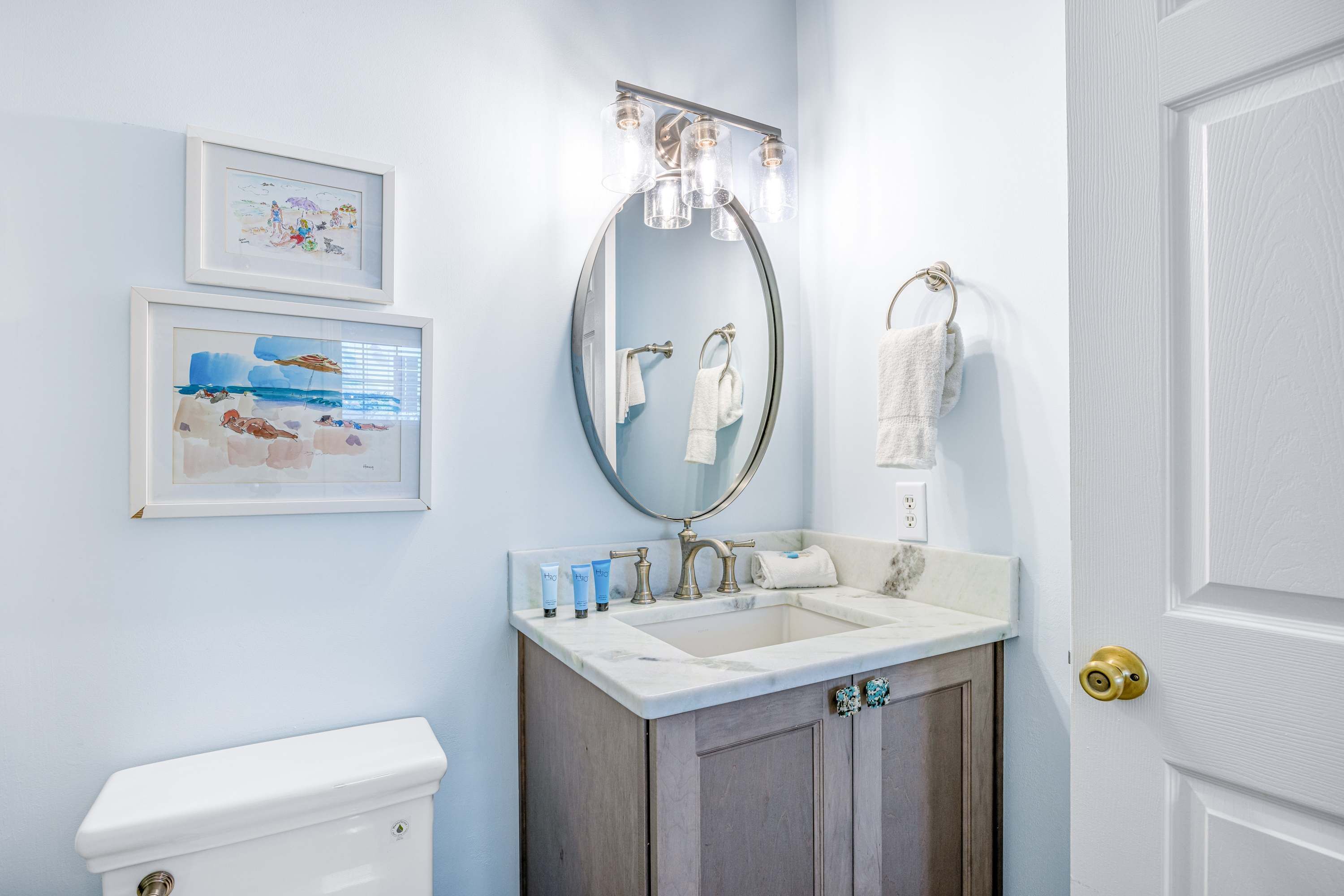 Ein Badezimmer mit Toilette, einem Waschbecken mit Marmorplatte, einem Spiegel mit Beleuchtung und gerahmten Kunstwerken an der Wand.
