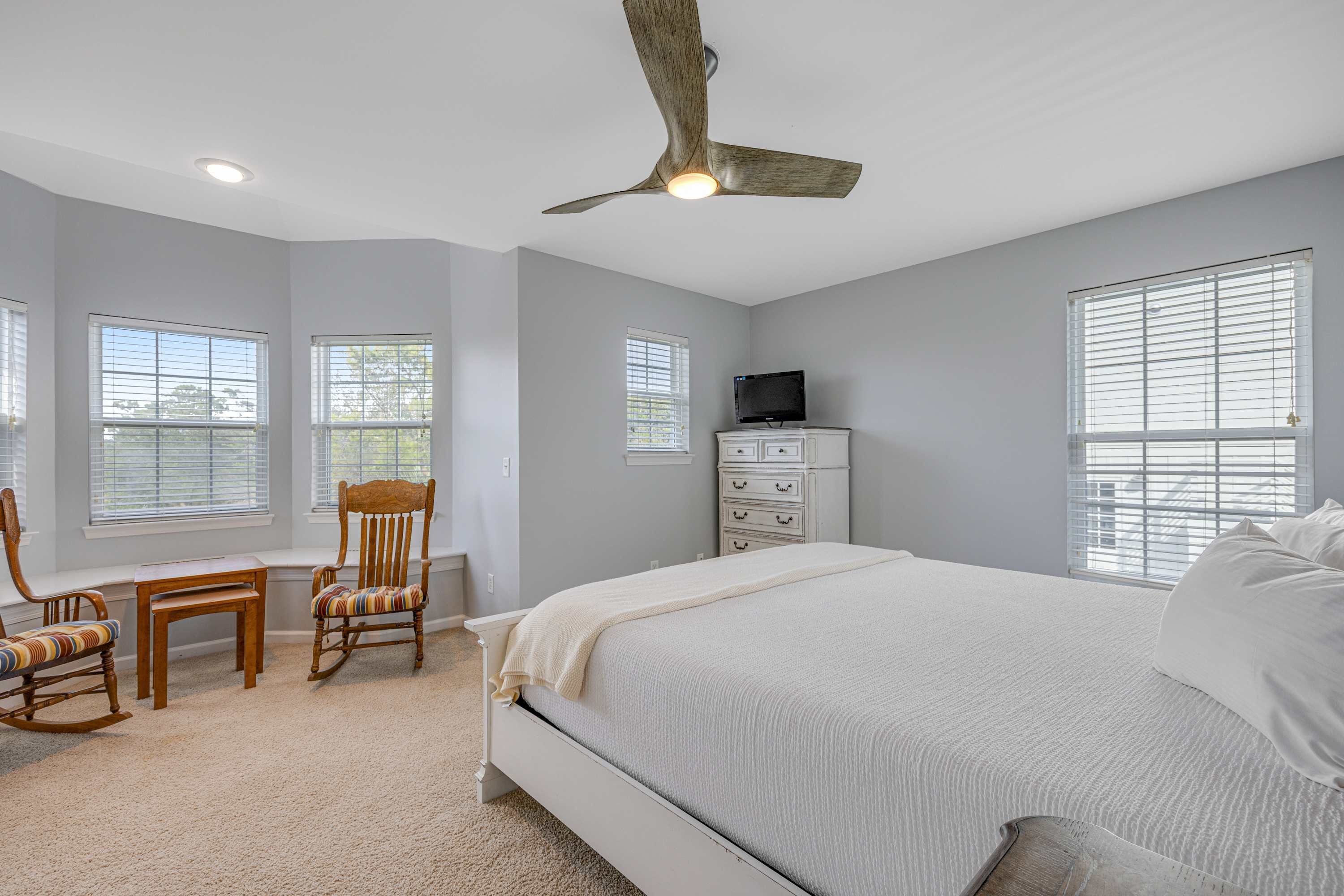 A bedroom with a large bed, a ceiling fan, a television on a dresser, and a window with blinds.