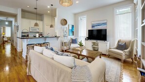 Una sala de estar moderna con un sofá, una mesa de centro y un televisor. Se observa una zona de cocina con armarios y un microondas.