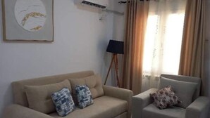 Olohuoneessa on beige sohva, pienempi nojatuoli, pyöreä sohvapöytä ja lattiavalaisin.