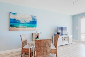 A dining area with wicker chairs, a round table, and a painting of a beach scene. - Snowbird-friendly beach condo with pools, putting green, shopping, & dining (Panama City Beach)