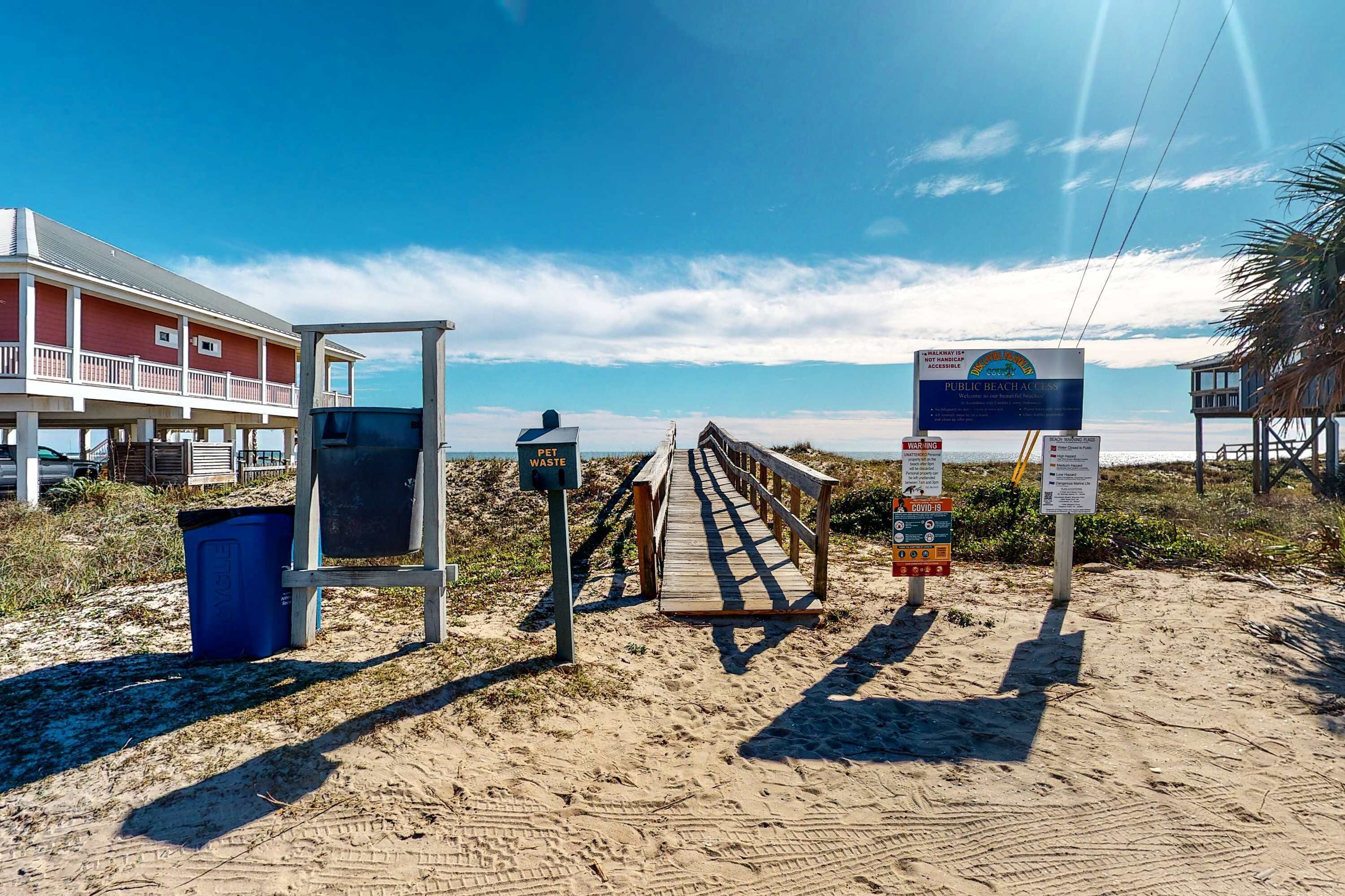 Una entrada a la playa con un pasillo de madera que conduce a un edificio con balcón. Se observan carteles que indican acceso público a la playa y disposición de desechos de mascotas.