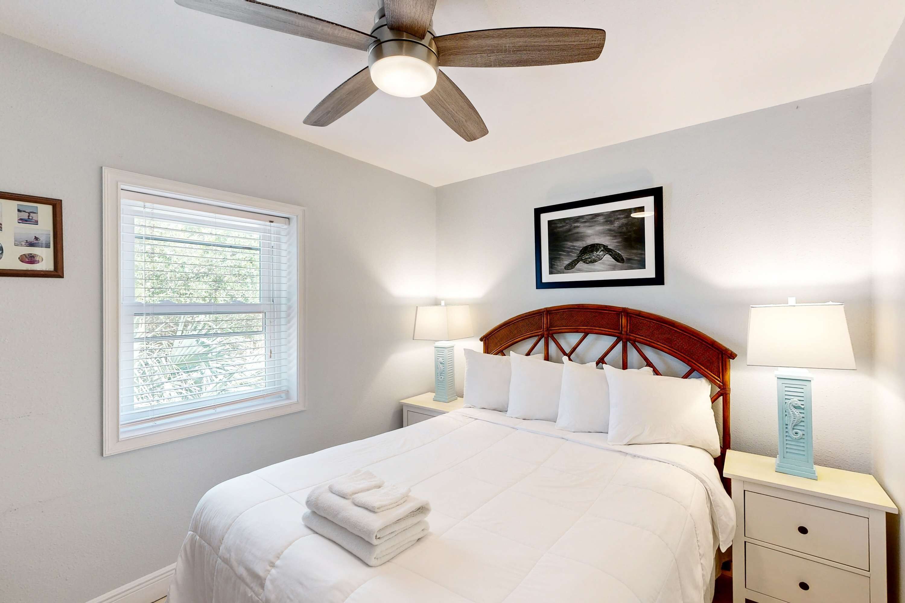 A neatly made bed with white linens and a folded towel, a ceiling fan, a window with blinds, two bedside lamps, and a framed picture on the wall.