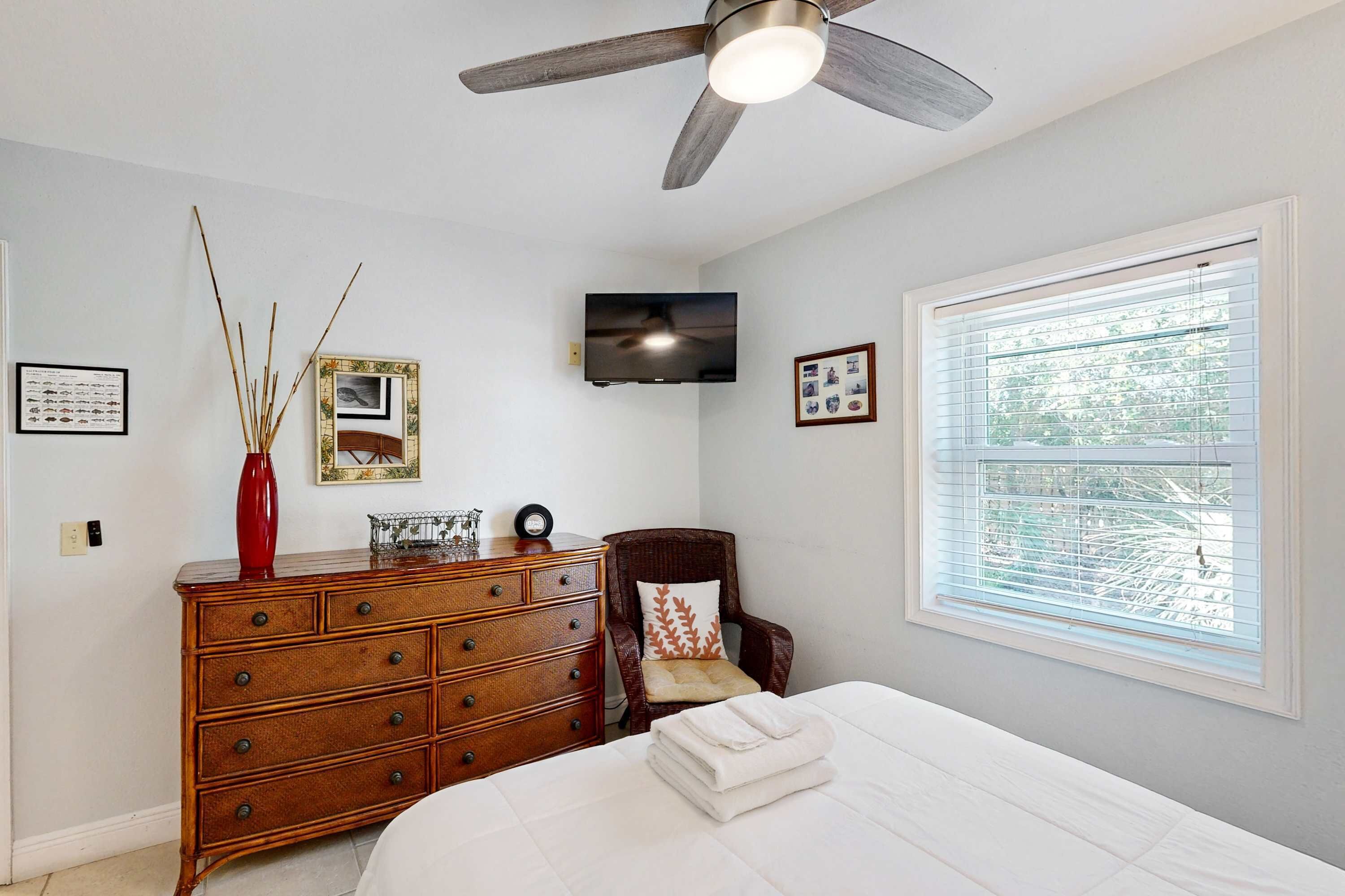 A bedroom with a bed, dresser, chair, ceiling fan, TV, and window with blinds.
