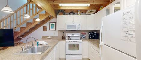 A modern kitchen with white cabinets, a stainless steel sink, a microwave, and a refrigerator. There is a wooden staircase with a view of the living room.