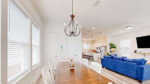 Un coin repas avec une table en bois, des chaises blanches et un lustre. On y trouve également un canapé bleu et un téléviseur fixé au mur.