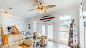 Uma sala de estar espaçosa com um ventilador de teto alto, escada de madeira e vista para o exterior por grandes janelas.