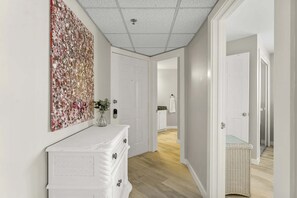 A hallway with a white dresser, a floral artwork on the wall, and a wicker basket. - Condo near the water with pool, tennis, balcony, grill, washer/dryer, & AC (Panama City Beach)