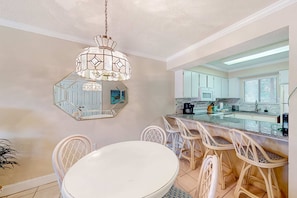 A dining area with a round table and white chairs, a decorative chandelier, a large mirror, and a kitchen with white cabinets and a countertop. - Dog-friendly resort condo with pools, hot tub, tennis, basketball, fitness room (Panama City Beach)