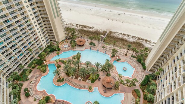 A high-rise hotel with a central swimming pool surrounded by palm trees and lounge areas, overlooking a beach. - Beachfront retreat with pool, hot tub, gym, & seasonal beach chairs & umbrella (Panama City Beach)