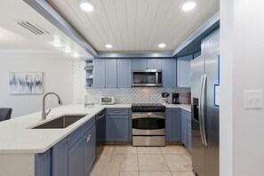 A modern kitchen with blue cabinets, stainless steel appliances, a herringbone tile backsplash, and recessed ceiling lights. - Beachfront Townhouse w/ Modern Upgrades Shared Pool, Fast WiFi, Outdoor Shower (Panama City Beach)