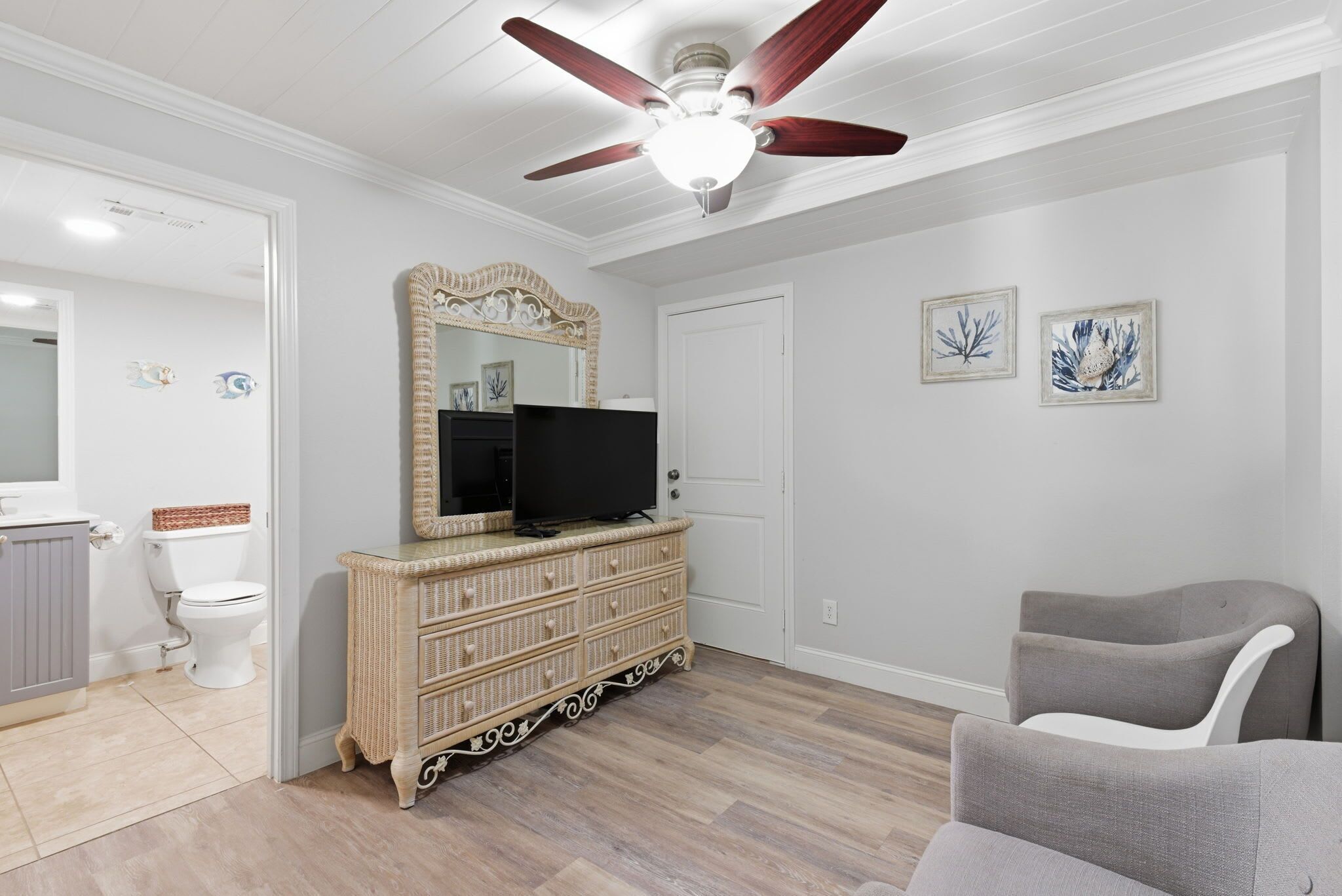 Una habitación con ventilador de techo, un televisor sobre un aparador de mimbre, un sillón gris y dos cuadros en la pared.