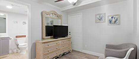 Una habitación con ventilador de techo, un televisor sobre un aparador de mimbre, un sillón gris y dos cuadros en la pared.